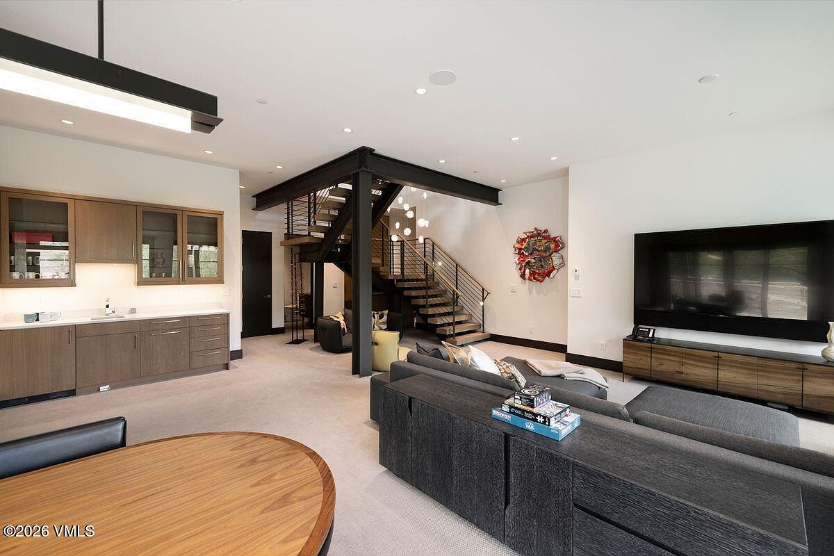 This spacious, open-concept living area features a contemporary design with a prominent industrial-style staircase as a central architectural focal point. The room includes a wet bar with wood cabinetry, a large sectional sofa facing a wall-mounted television, and a dining area in the foreground. The neutral color palette and clean lines create a sophisticated, modern atmosphere suitable for entertaining.