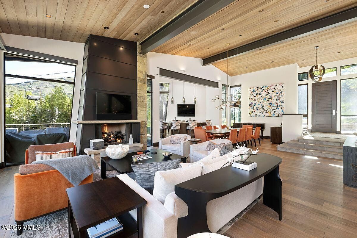 This expansive open-concept living area features a striking dark metal-clad chimney breast as a central focal point, complemented by a warm wood-paneled ceiling. The space is furnished with a plush light-toned sofa, a dark coffee table, and vibrant orange accent chairs, all set against a backdrop of large windows that frame mountain views. A dining area with a long table and modern lighting fixtures is visible in the background, creating a cohesive and sophisticated mountain-modern aesthetic.