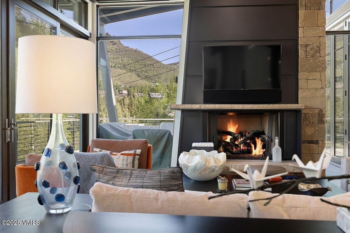 This cozy living room features a prominent fireplace with a dark, modern surround and a stone mantel, creating a warm focal point. Large windows provide an expansive view of a mountain landscape with a ski lift visible in the distance. The space is furnished with comfortable seating, including an orange armchair and a plush sofa, accented by a decorative glass lamp and a unique sculptural bowl on the coffee table.