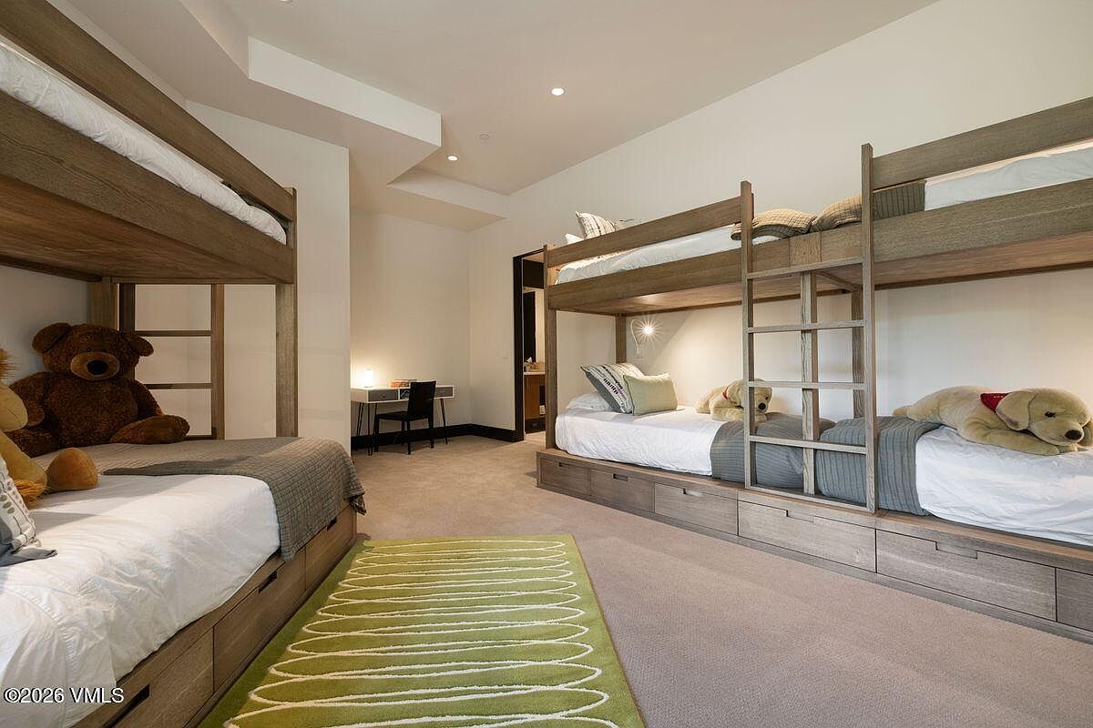 This spacious bedroom features two sets of custom light wood bunk beds, providing ample sleeping arrangements. A green patterned area rug sits on the carpeted floor, while a small desk and chair are tucked into the corner near a doorway. The room is brightly lit with recessed lighting and offers a clean, functional design suitable for a family home.