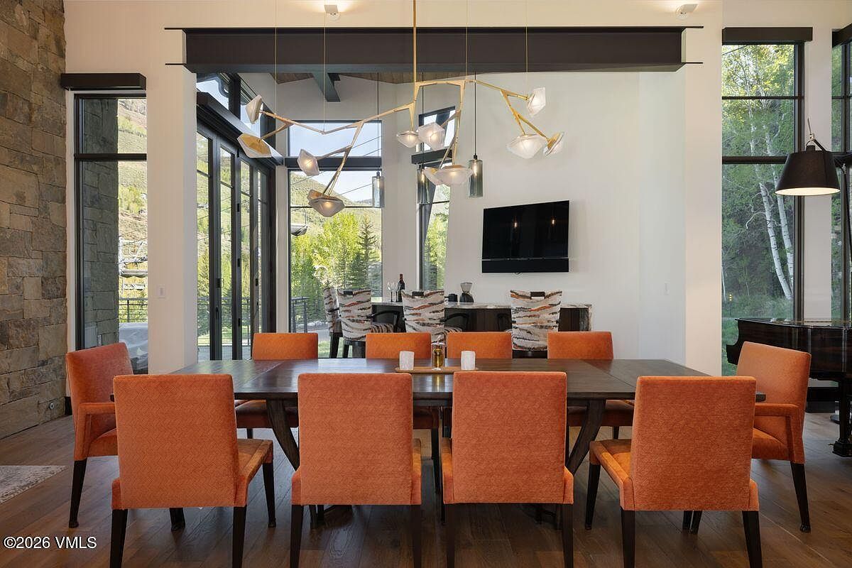 This elegant dining area features a large dark wood table surrounded by eight vibrant orange upholstered chairs, creating a bold focal point. Above the table hangs a modern, multi-armed gold chandelier, while the background showcases a bar area with stools and a television mounted on a white wall. The room is framed by large windows that offer expansive views of the surrounding trees and landscape, enhancing the bright and airy atmosphere.