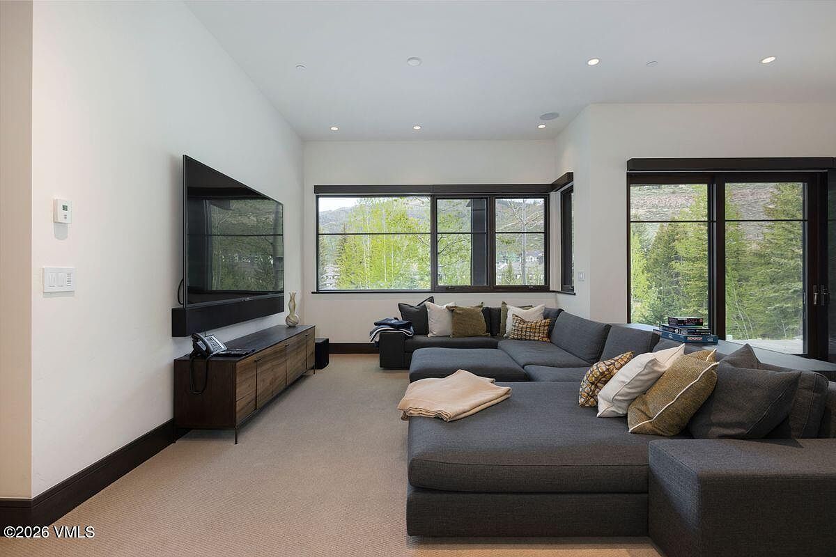 This contemporary living room features a large, dark gray sectional sofa arranged for comfortable viewing of a wall-mounted television. A long, dark wood media console sits beneath the television, while large windows provide ample natural light and scenic views of the surrounding trees and mountains. The space is finished with neutral carpeting and recessed lighting, creating a clean and inviting atmosphere.
