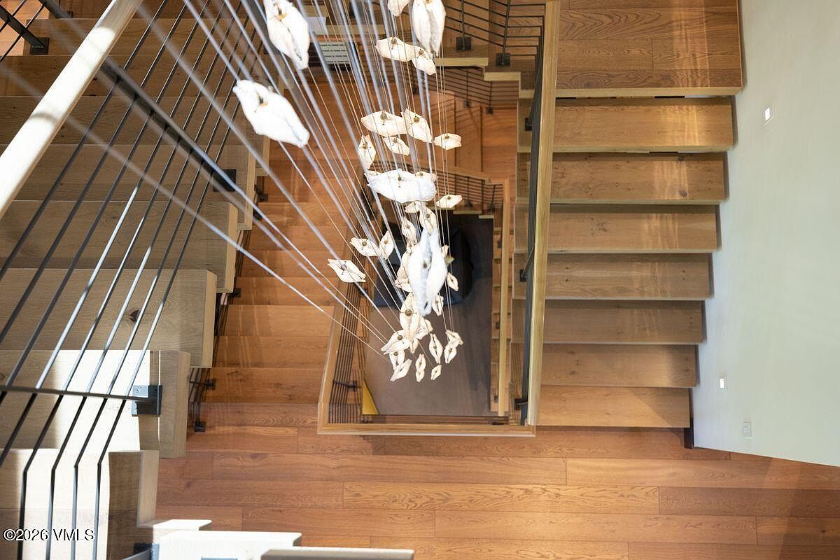 This high-angle, top-down perspective captures a dramatic staircase design featuring warm-toned wood treads and a sleek, dark metal railing. A striking, multi-tiered chandelier with numerous organic-shaped, translucent pendants hangs centrally, creating a focal point that draws the eye through the open stairwell. The composition emphasizes the architectural geometry of the staircase and the interplay of light and shadow across the wooden surfaces.