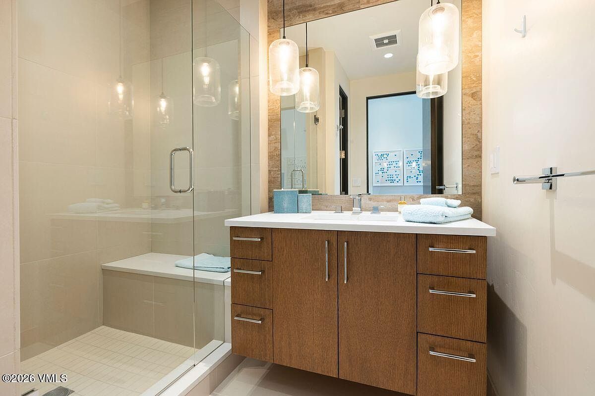 This modern bathroom features a wood-toned vanity with a white countertop and a large, rectangular mirror framed by a stone-tiled wall. To the left, a glass-enclosed walk-in shower includes a built-in bench and light-colored wall tiles. The space is illuminated by two contemporary pendant lights hanging above the vanity, creating a clean and functional aesthetic.