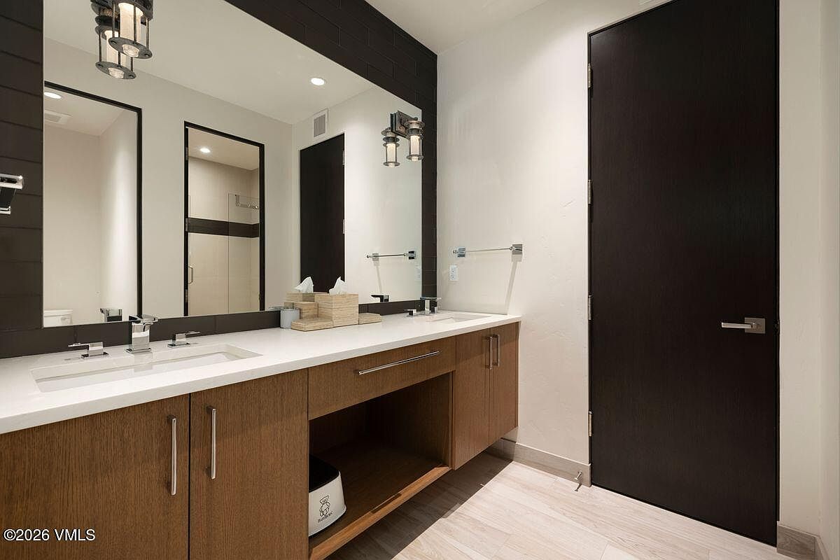 This modern bathroom features a double vanity with a white countertop and light wood cabinetry, accented by a dark, textured wall behind the large mirror. The space is illuminated by two wall-mounted light fixtures and recessed lighting, creating a clean and contemporary aesthetic. A dark, full-height door provides a striking contrast to the neutral walls and light-toned flooring.