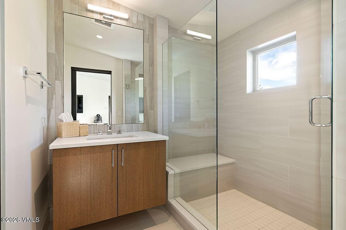 This modern bathroom features a sleek vanity with a light wood cabinet and a white countertop, paired with a large, frameless mirror. To the right, a glass-enclosed walk-in shower includes a built-in bench and light-toned wall tiles. The space is bright and minimalist, offering a clean, contemporary aesthetic.