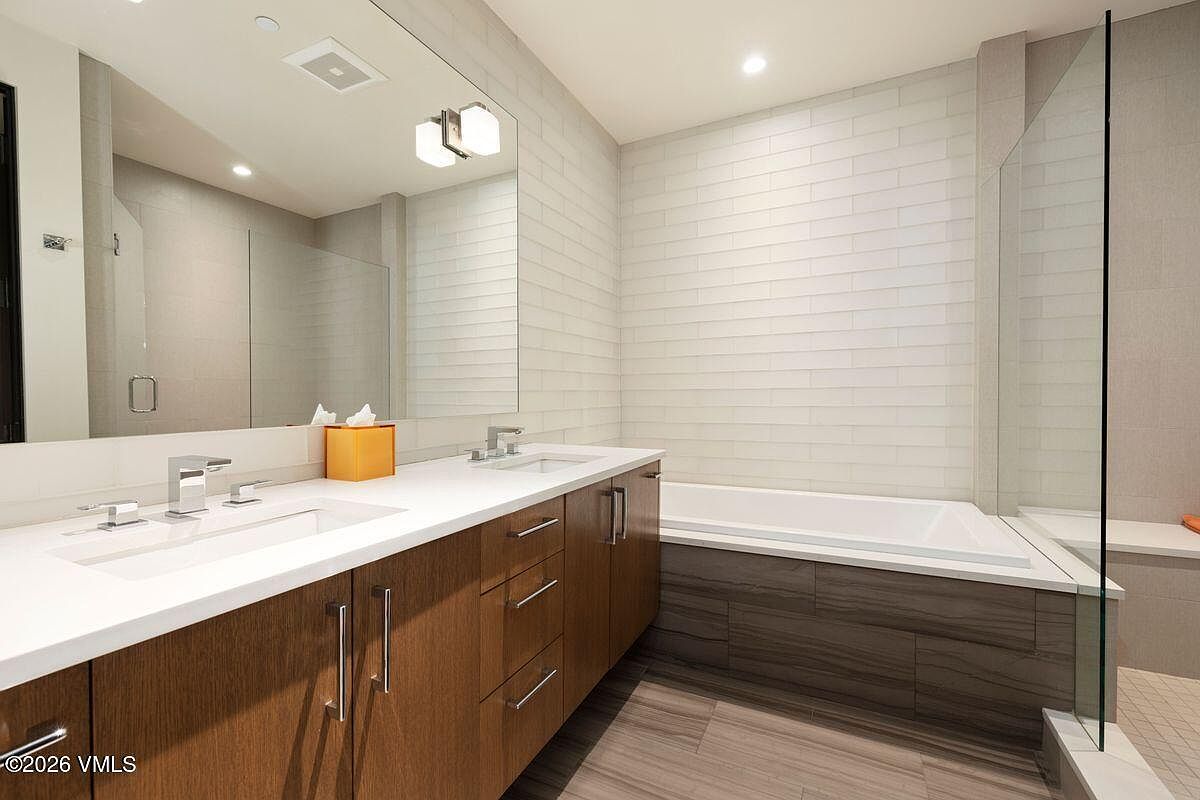 This modern bathroom features a double vanity with dark wood cabinetry and a white countertop, complemented by a large wall-mounted mirror. To the right, a deep soaking tub is set against a wall of light-colored rectangular tiles, adjacent to a glass-enclosed shower area. The space is finished with wood-look tile flooring, creating a clean and contemporary aesthetic.
