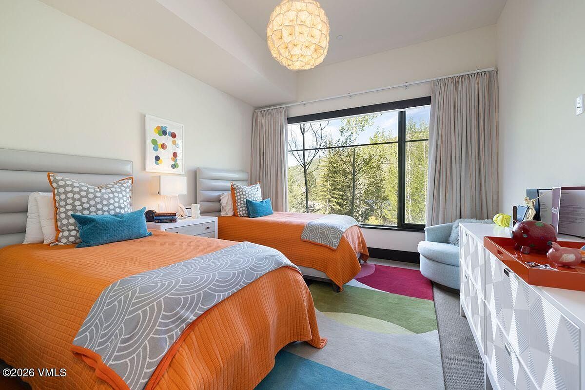 This bright and cheerful guest bedroom features two twin beds with vibrant orange bedding and patterned grey accents. A large window provides ample natural light and a scenic view of the surrounding trees, while a colorful area rug adds a playful touch to the space. The room is completed by a white dresser, a light blue armchair, and a unique textured pendant light fixture.