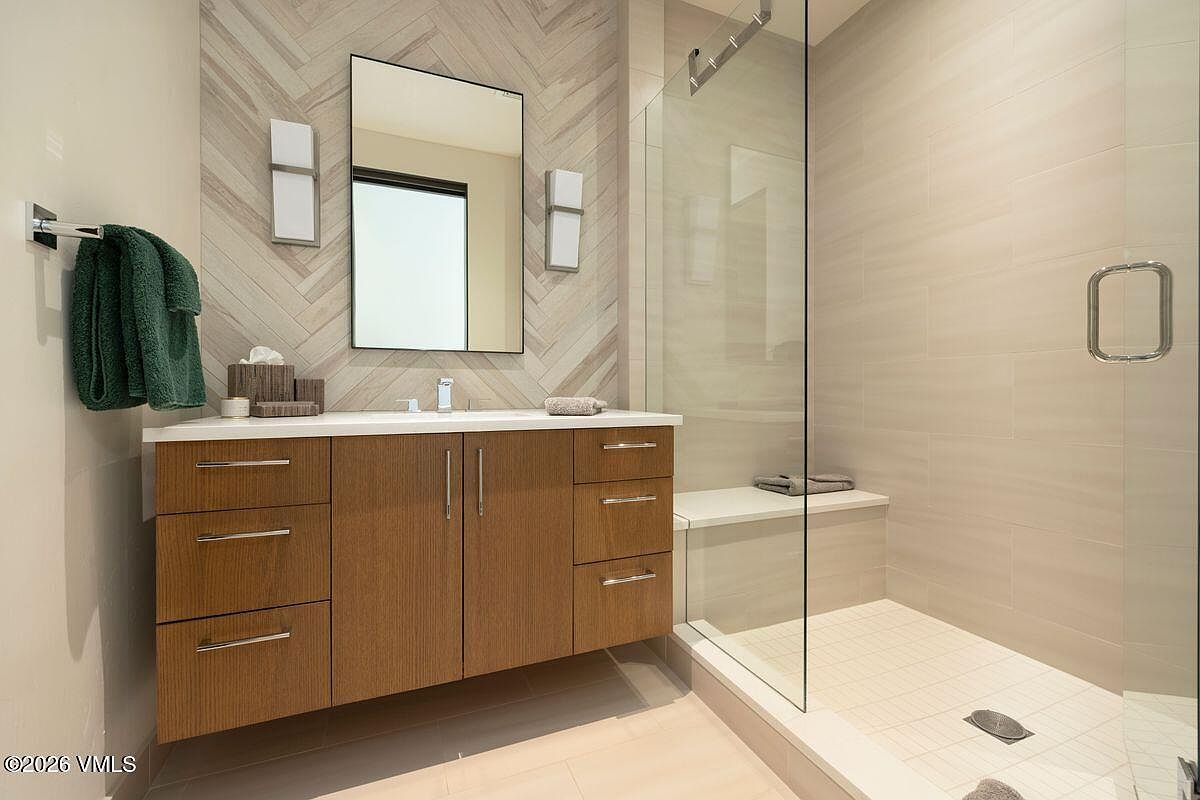 This modern bathroom features a sleek vanity with light wood cabinetry and a white countertop, complemented by a rectangular mirror flanked by two wall-mounted light fixtures. A glass-enclosed walk-in shower with a built-in bench and light-toned wall tile adds a spacious, spa-like feel to the room. The wall behind the vanity is accented with a herringbone-patterned tile, creating a sophisticated focal point in this contemporary space.