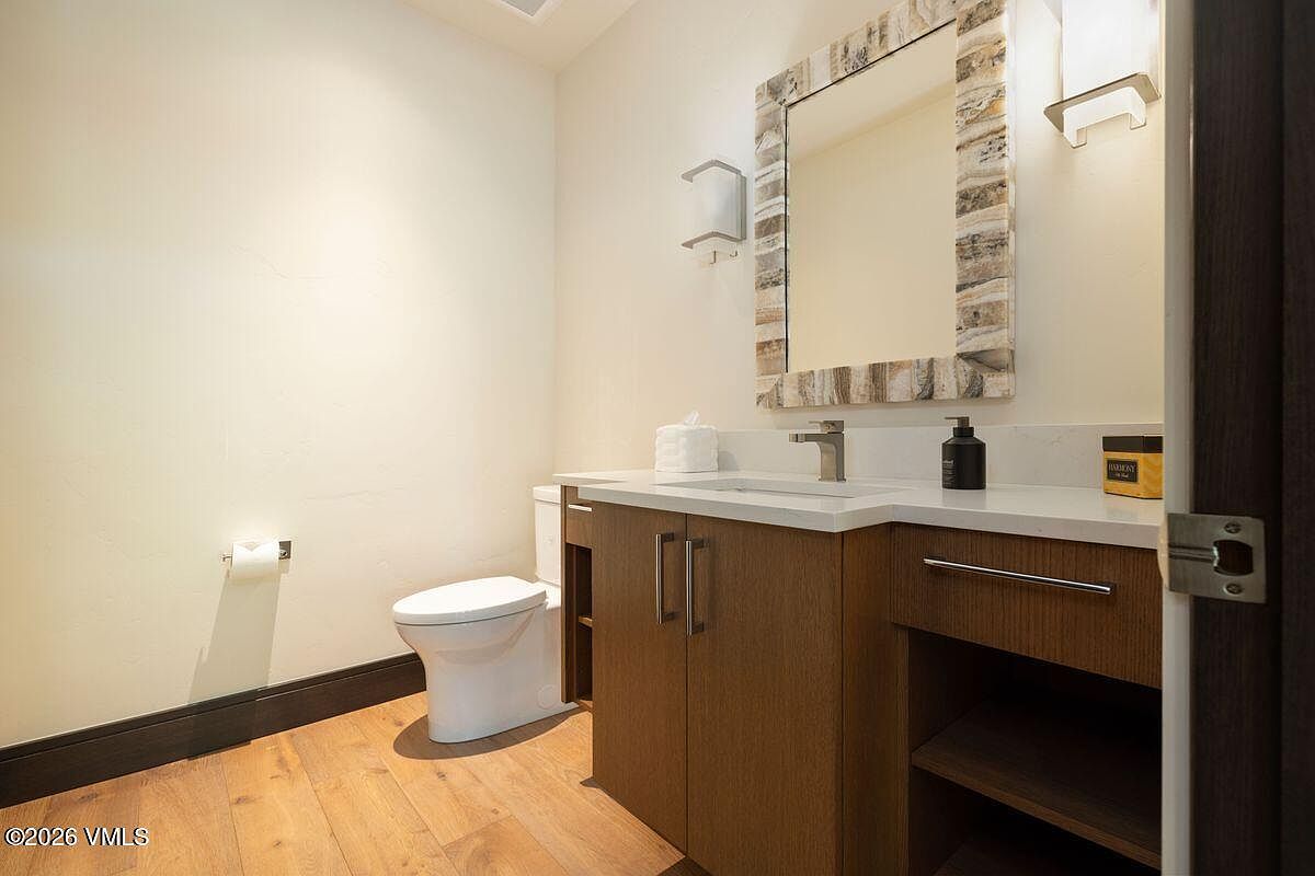 This modern powder room features a sleek vanity with a light-toned countertop and a rectangular undermount sink, complemented by a decorative stone-tiled mirror frame. The space includes a white toilet, light-colored walls, and warm-toned wood-look flooring, creating a clean and contemporary aesthetic. The perspective is a straight-on shot capturing the vanity and toilet area, highlighting the efficient use of space.