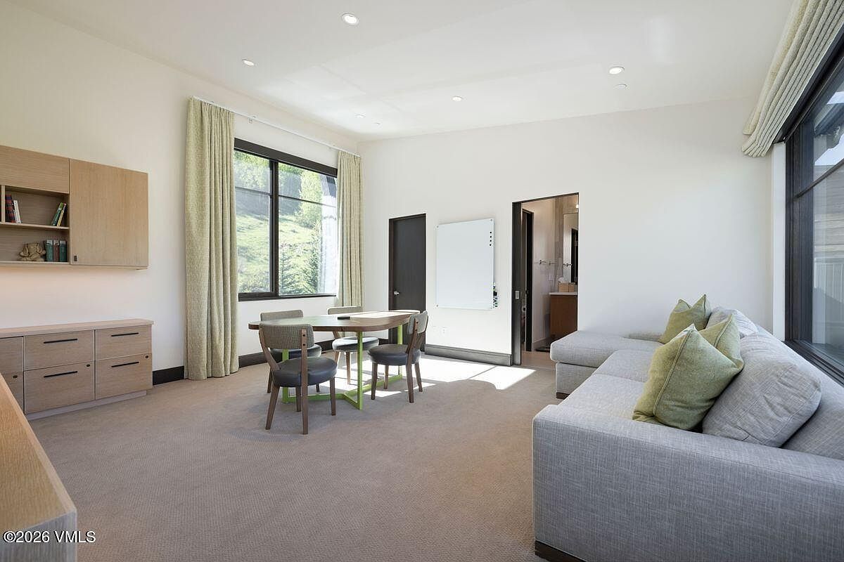 This versatile room functions as a modern home office or study, featuring a light-toned desk with chairs and a comfortable gray sectional sofa. The space is brightened by large windows with light-colored curtains and includes built-in cabinetry for storage alongside a wall-mounted whiteboard. The neutral color palette and clean lines create a professional yet inviting atmosphere suitable for work or relaxation.