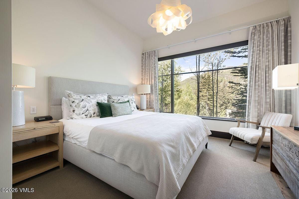 This serene primary bedroom features a neutral color palette with a plush upholstered bed frame and layered bedding. A large window wall provides expansive views of the surrounding forest and mountains, while a modern sculptural light fixture hangs centrally above the bed. The room is completed by light wood nightstands and a comfortable armchair, creating a tranquil and sophisticated retreat.