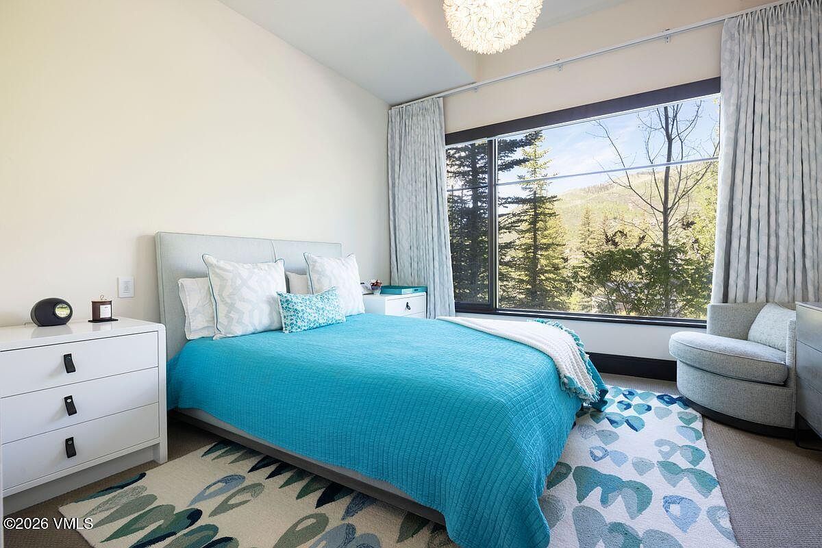This bright and inviting bedroom features a queen-sized bed with a vibrant blue quilt and patterned accent pillows. A large window provides an expansive view of the surrounding forest and mountains, while a patterned area rug adds texture to the space. The room is completed by a white dresser, a modern swivel chair, and a unique textured pendant light fixture.