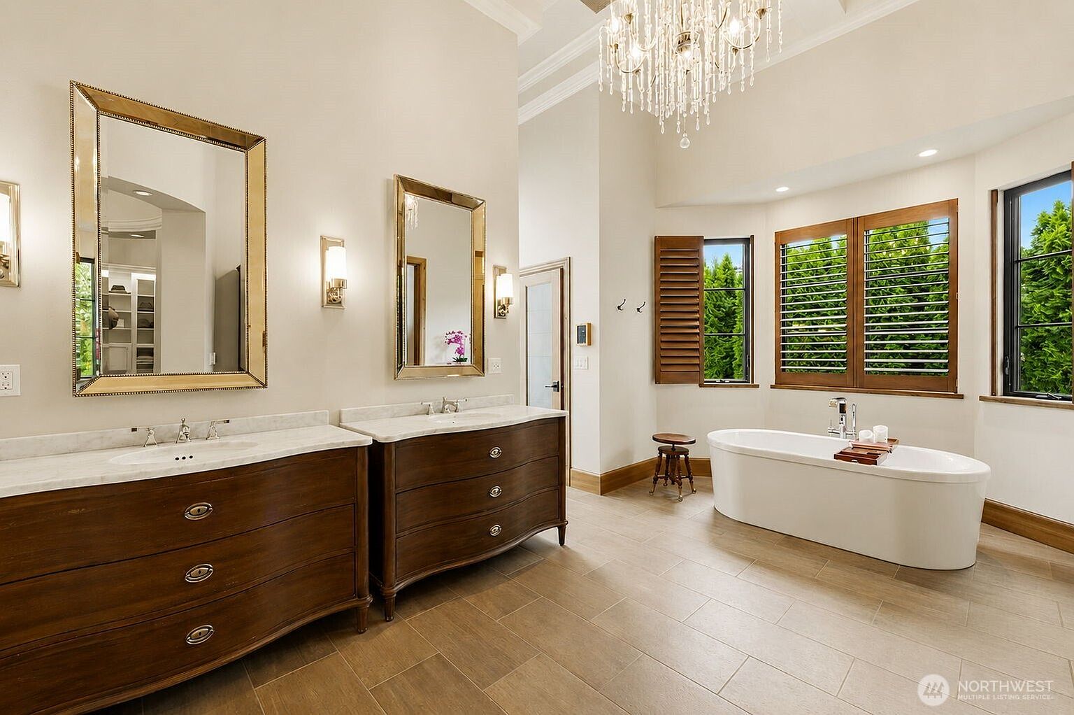 This spacious primary bathroom features a double vanity with dark wood cabinetry and light stone countertops, complemented by two large, framed mirrors. A freestanding soaking tub sits near a set of windows with wooden shutters, while a crystal chandelier hangs from the high ceiling, adding a touch of elegance. The room is finished with neutral-toned tile flooring, creating a bright and sophisticated atmosphere.