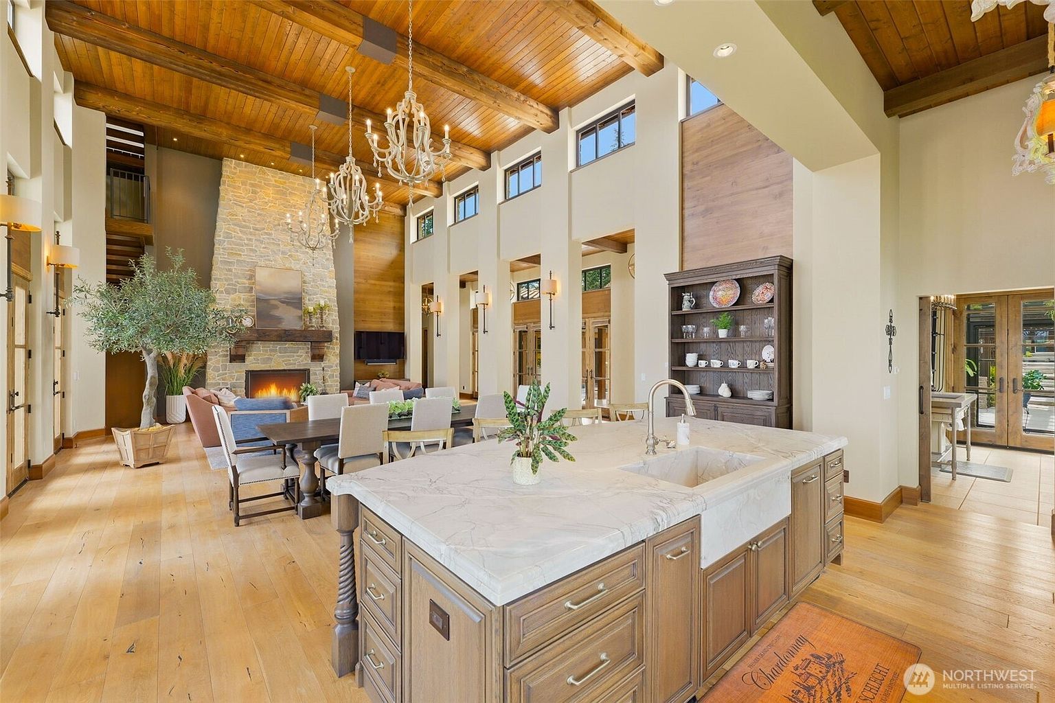 This expansive open-concept living space features a large kitchen island with a stone countertop and a farmhouse sink, seamlessly flowing into a dining and living area. The room is defined by a high, vaulted wood-paneled ceiling with exposed beams and a prominent stone-clad chimney breast housing a fireplace. Large windows and chandeliers provide ample light, creating an airy, sophisticated atmosphere with a blend of rustic and refined design elements.