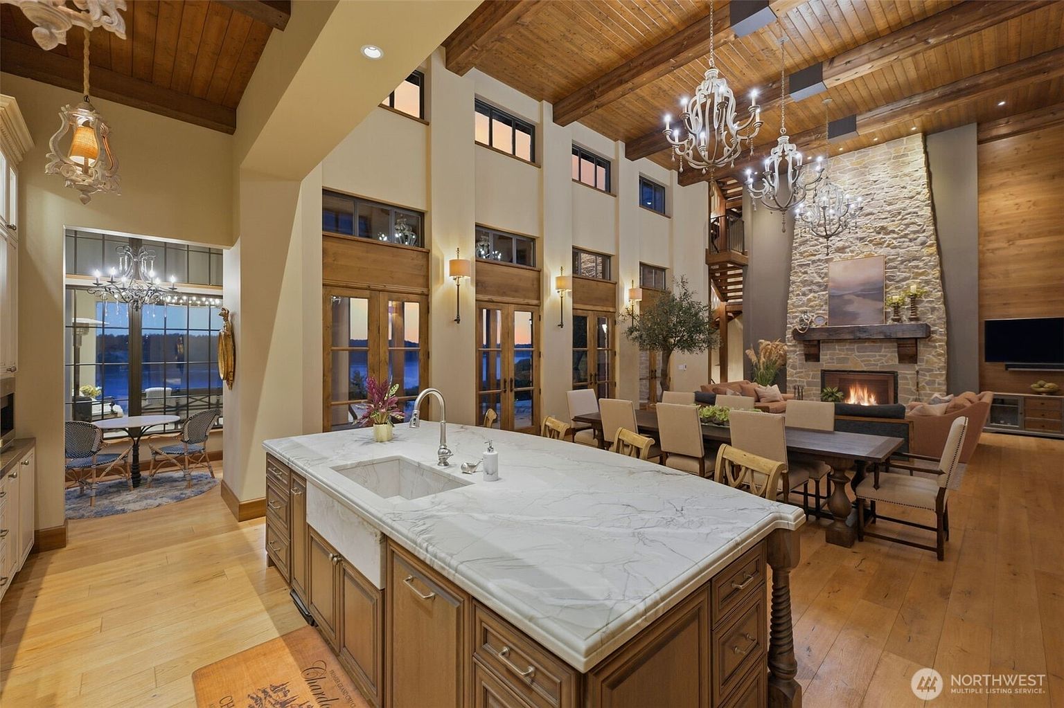 This expansive open-concept kitchen and dining area features a large central island with a stone countertop and a farmhouse-style sink. The room is defined by its high wood-paneled ceiling with exposed beams, a prominent stone-clad chimney breast housing a fireplace, and multiple sets of tall glass doors that provide views of the water. The space is elegantly lit by ornate chandeliers and wall sconces, creating a warm and inviting atmosphere suitable for both cooking and formal dining.