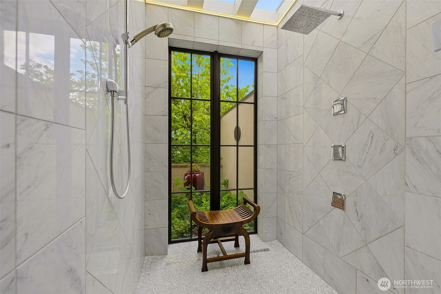 This luxurious walk-in shower features light-colored stone wall tiles arranged in a diamond pattern and a textured stone floor. A tall, black-framed window provides a view into a private garden, while a wooden stool sits in the center. The space is equipped with both a ceiling-mounted rainfall showerhead and a wall-mounted handheld sprayer, creating a spa-like atmosphere.