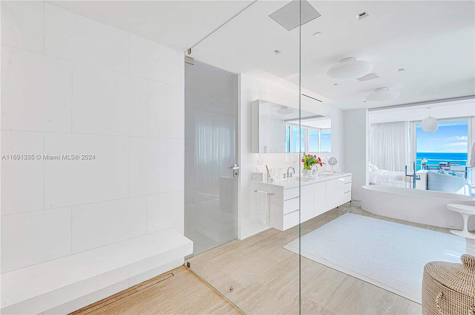This is a bright and luxurious primary bathroom featuring a modern design with white walls, cabinetry, and fixtures. A glass-enclosed shower and a freestanding bathtub offer spa-like amenities, while a large vanity with dual sinks provides ample space. The bathroom opens to a bedroom area with ocean views, enhancing the sense of tranquility and luxury.