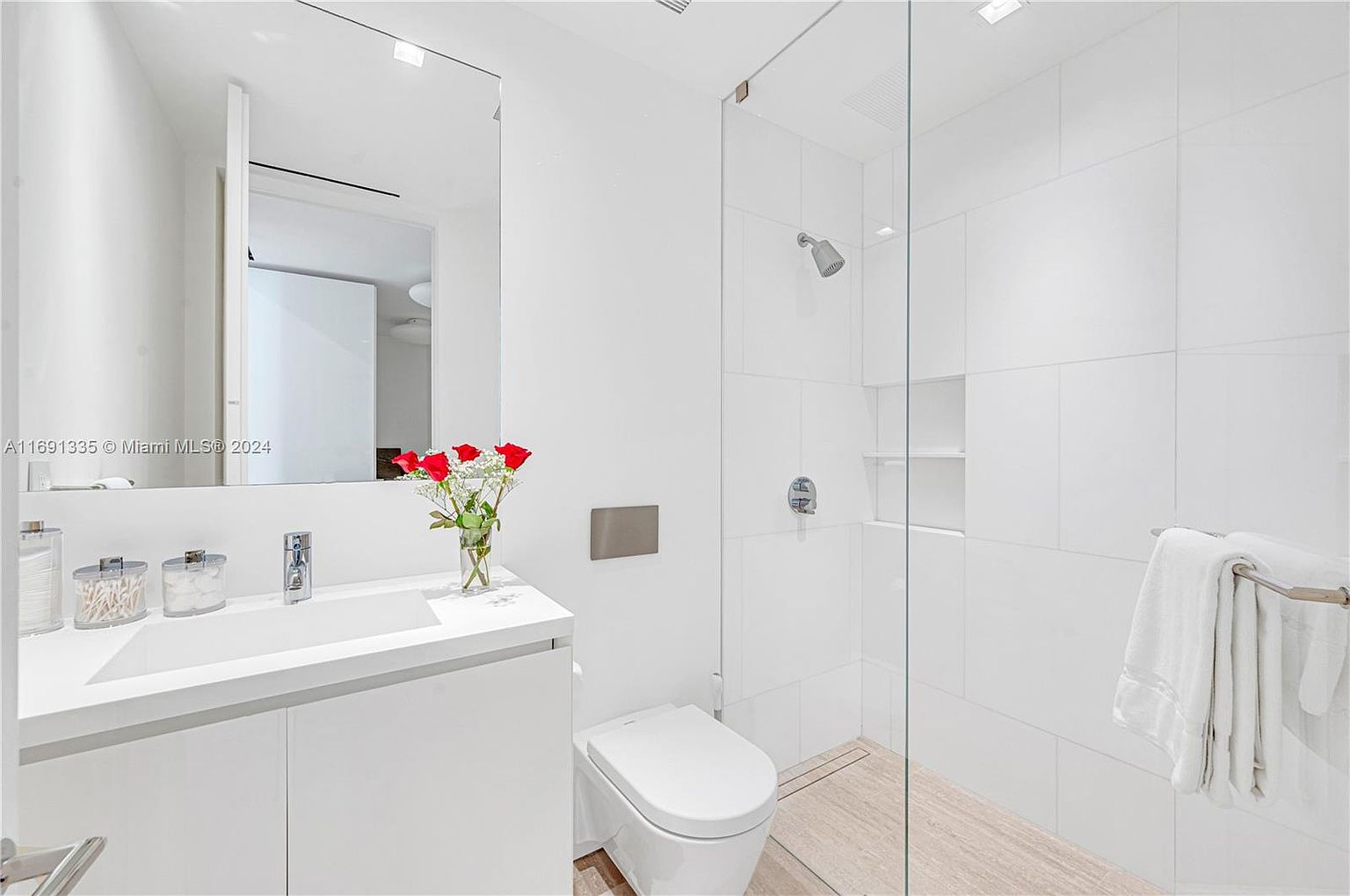 This is a bright and modern primary bathroom featuring a sleek, white vanity with a rectangular sink and chrome faucet. A large mirror reflects the space, while a glass-enclosed shower with white tile and built-in shelving adds a luxurious touch. The toilet is a modern, low-profile design, and a vase of red roses adds a pop of color.