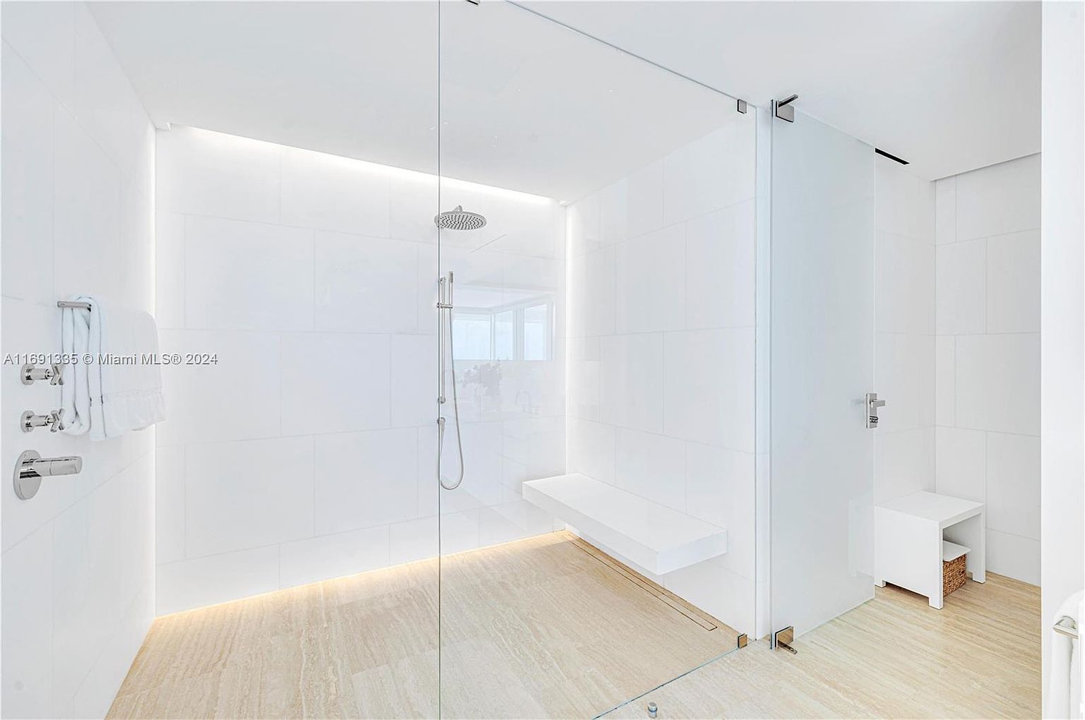 This is a bright and modern primary bathroom featuring a walk-in shower with glass enclosure, a built-in bench, and a separate stool. The walls are tiled in white, and the floor has a light-colored stone finish. The design is minimalist and clean, creating a spa-like atmosphere.