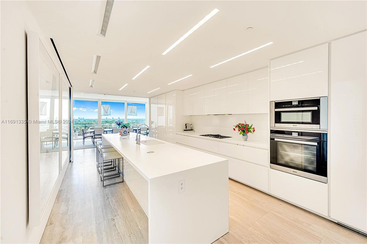 This is a bright, modern kitchen featuring sleek white cabinetry and a large central island with seating. Stainless steel appliances are integrated into the design, and large windows offer a view of the city skyline. The perspective is from a wide angle, showcasing the spaciousness and clean lines of the kitchen.