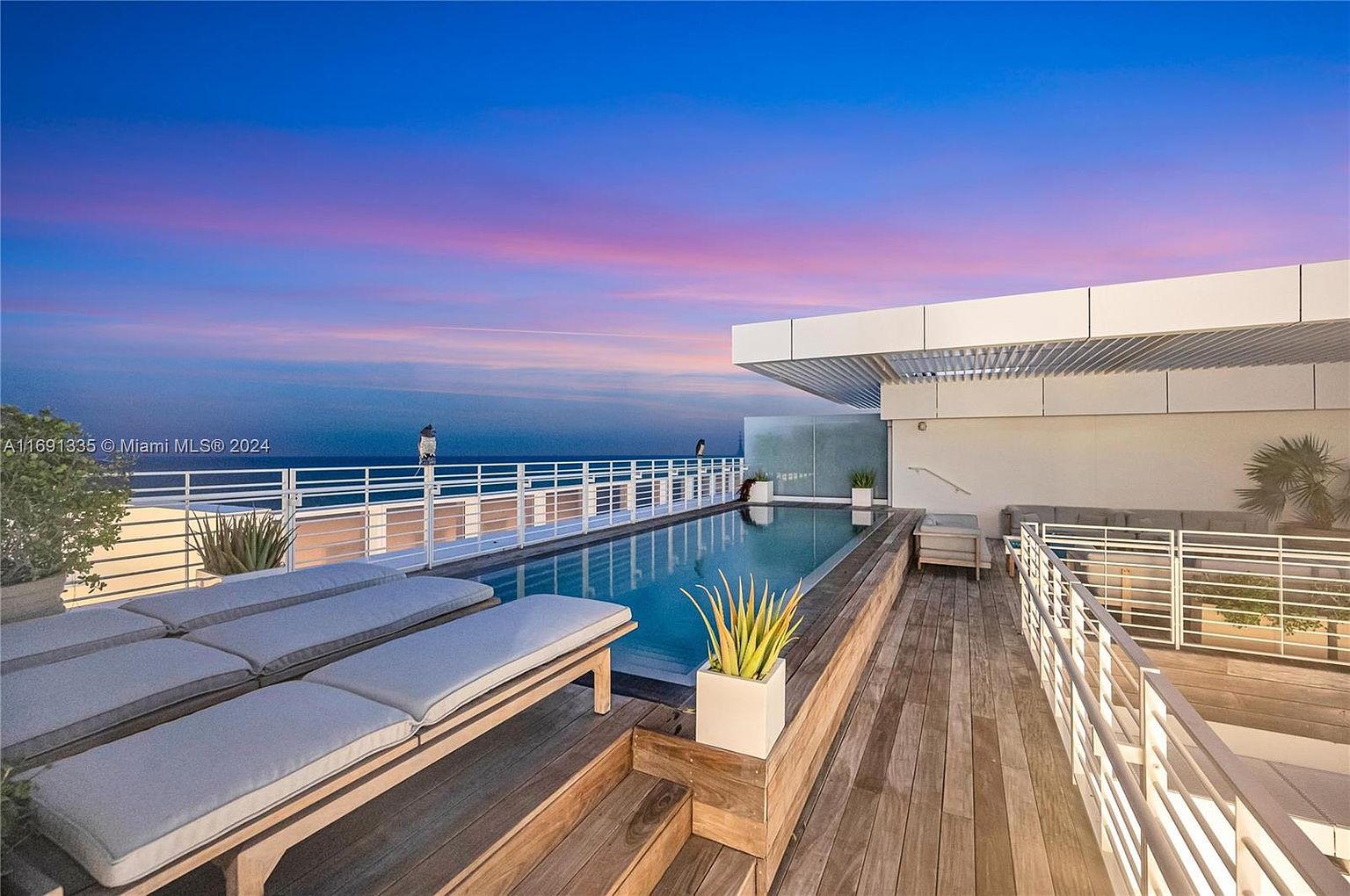 This image showcases a luxurious rooftop pool and deck area, perfect for relaxation and entertainment. The pool is surrounded by wooden decking, lounge chairs, and potted plants, creating a serene atmosphere. A modern building structure with white accents provides a stylish backdrop, while the ocean view adds to the overall appeal.