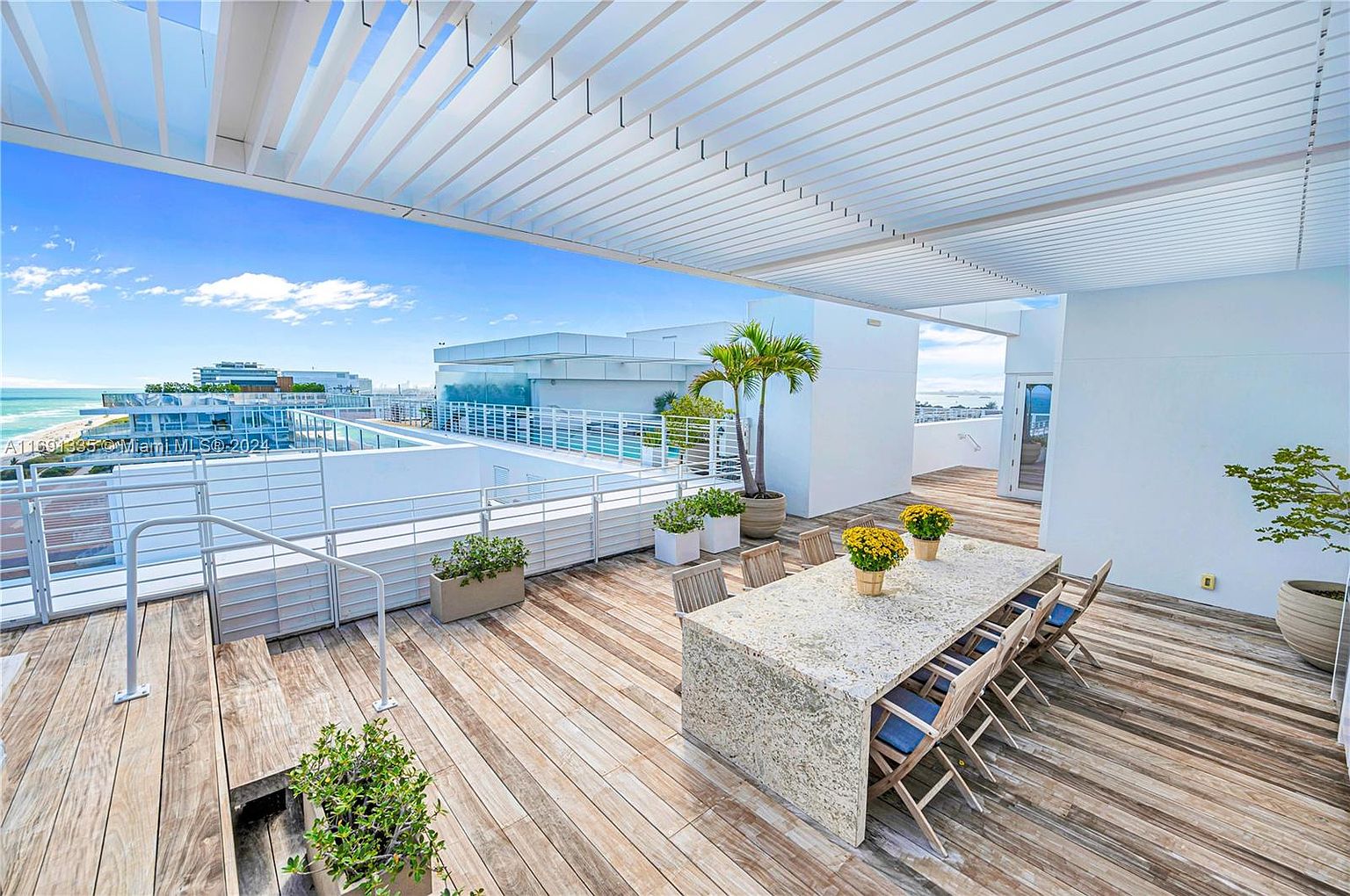 This image showcases a luxurious rooftop patio/deck/balcony with stunning ocean views. The space features a large dining table with seating, potted plants, and a modern pergola providing shade. The wooden decking adds warmth, while the white railings and building elements contribute to a clean, contemporary aesthetic, perfect for outdoor entertaining.