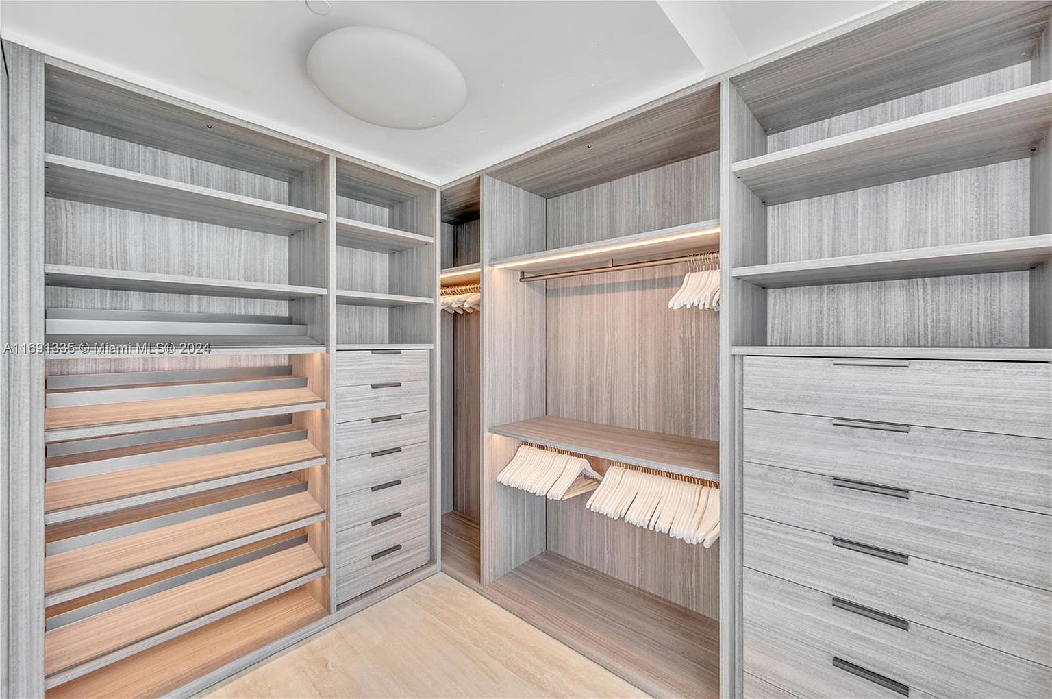 This is a well-organized walk-in closet featuring custom shelving, drawers, and hanging rods. The closet is finished in a light gray wood grain, providing a modern and clean aesthetic. The space is illuminated by built-in lighting, highlighting the ample storage and functionality.