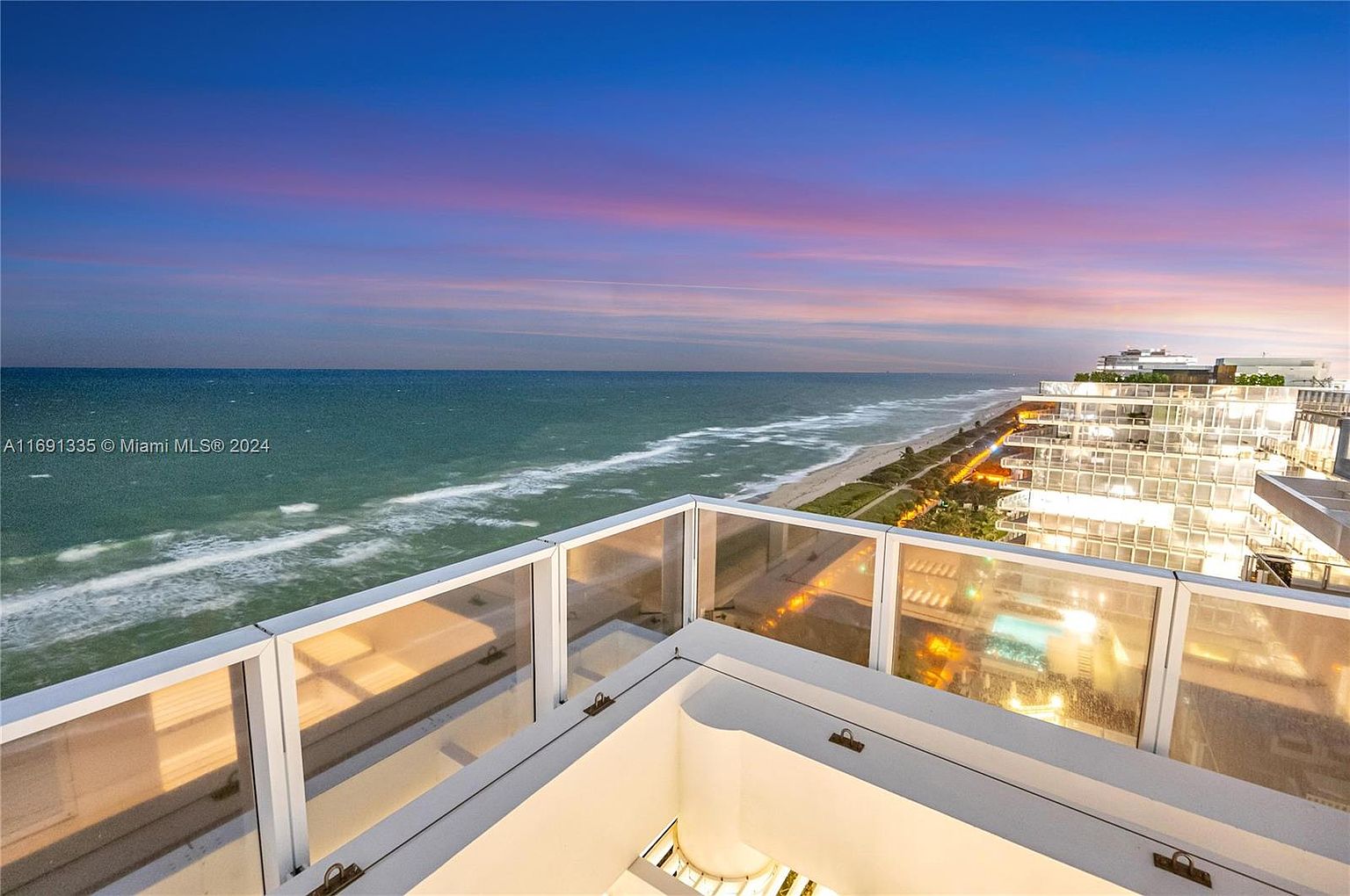 This image showcases a luxurious balcony view overlooking the ocean at sunset. The balcony features modern glass railings and a clean, white structure. In the background, the ocean meets the shoreline, with a high-rise building visible along the coast, creating a sense of upscale coastal living.