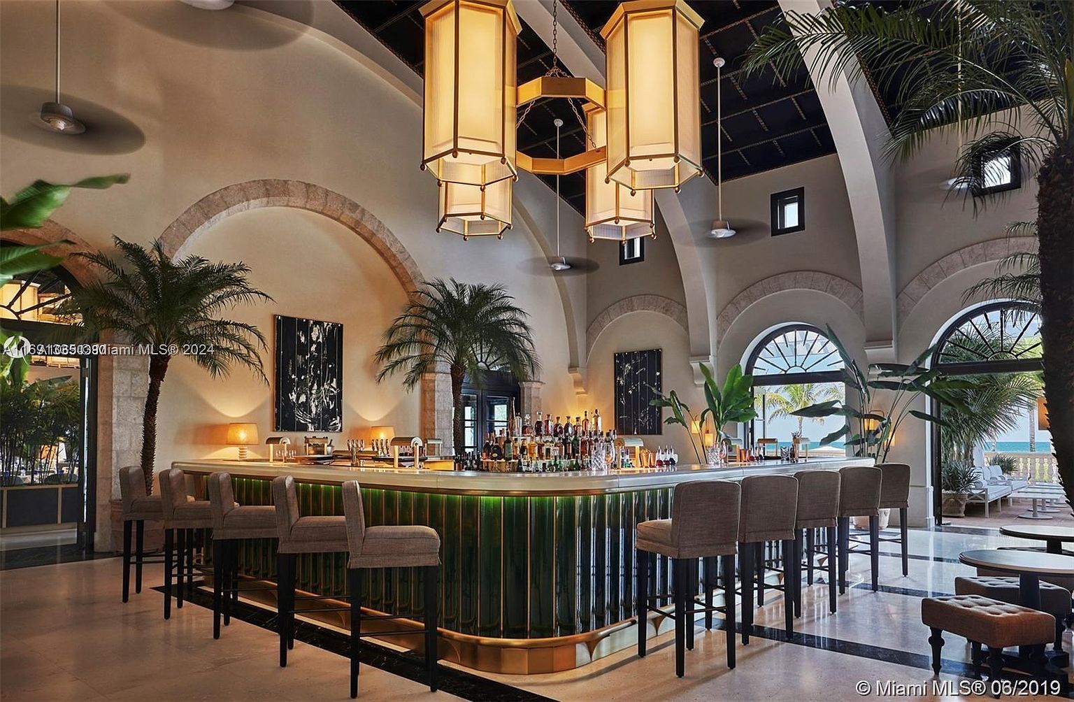 This interior shot showcases a luxurious bar area with a curved, illuminated counter, complemented by stylish bar stools. The space is adorned with tropical plants and elegant hanging light fixtures, creating a sophisticated and inviting atmosphere. Arched doorways and views of the ocean enhance the upscale ambiance, making it an ideal setting for relaxation and entertainment.