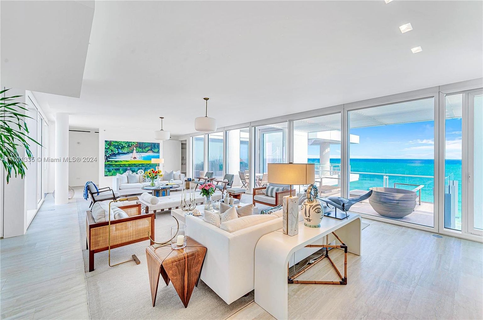 This is a bright and airy living room with floor-to-ceiling windows offering stunning ocean views. The space is furnished with modern white sofas, rattan chairs, and unique side tables, creating a sophisticated and comfortable atmosphere. The open layout and natural light enhance the sense of luxury and spaciousness.