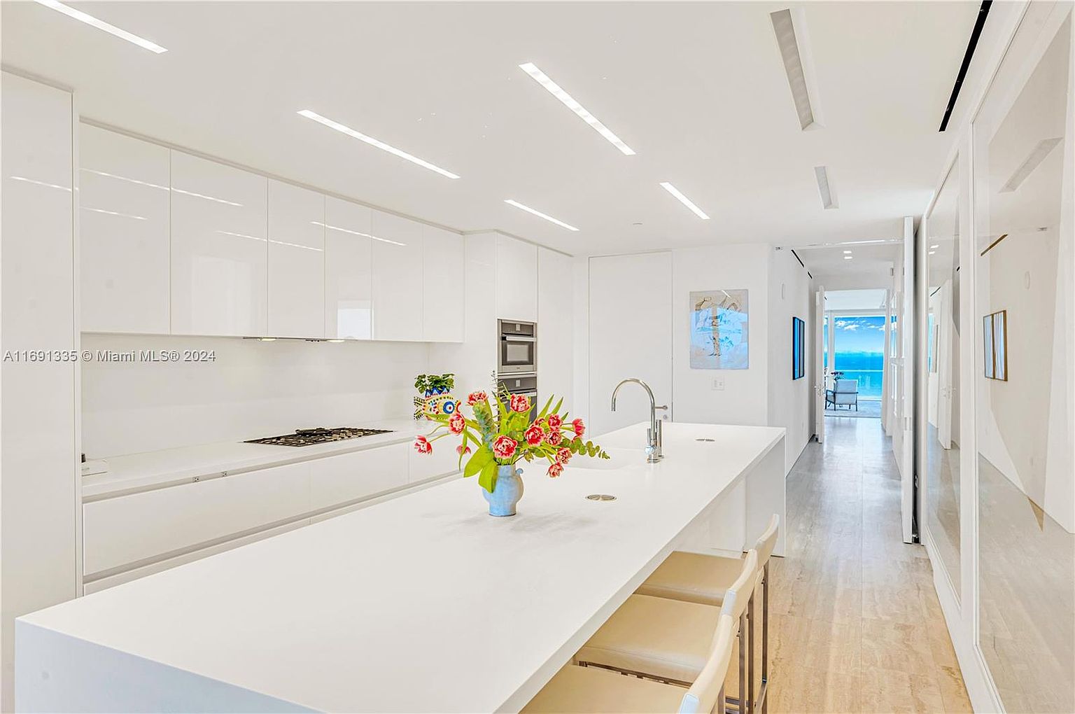 This is a bright, modern kitchen featuring sleek white cabinetry and countertops. A large island dominates the center of the room, adorned with a vase of flowers. The kitchen opens to a hallway with a view of the ocean, creating a sense of spaciousness and luxury.