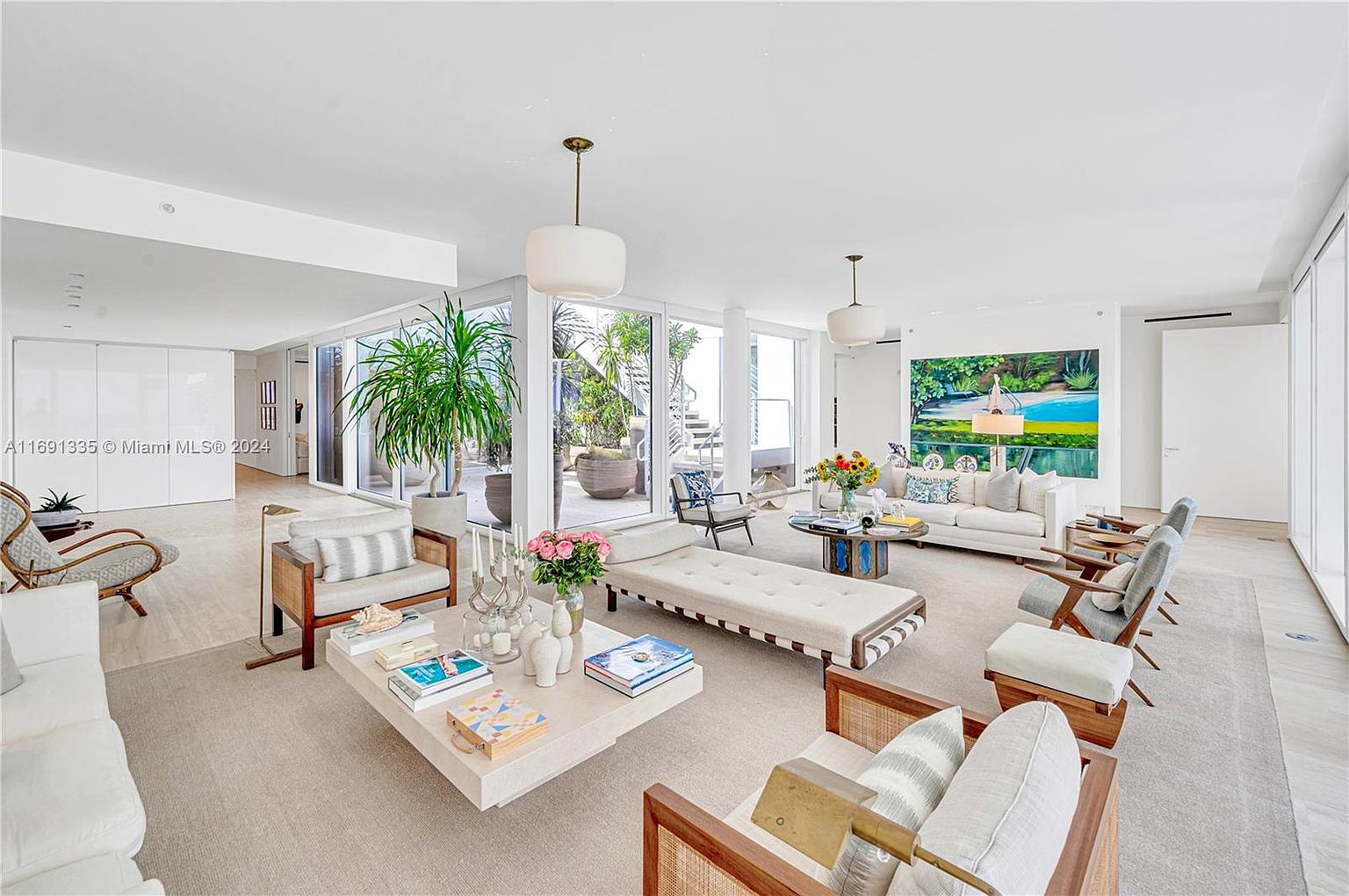 This is a bright and spacious living room featuring a neutral color palette with white walls and light-colored flooring. The room is furnished with modern furniture, including sofas, chairs, and a large coffee table. Large windows and glass doors provide ample natural light and views of the outdoor area, creating an open and airy atmosphere.