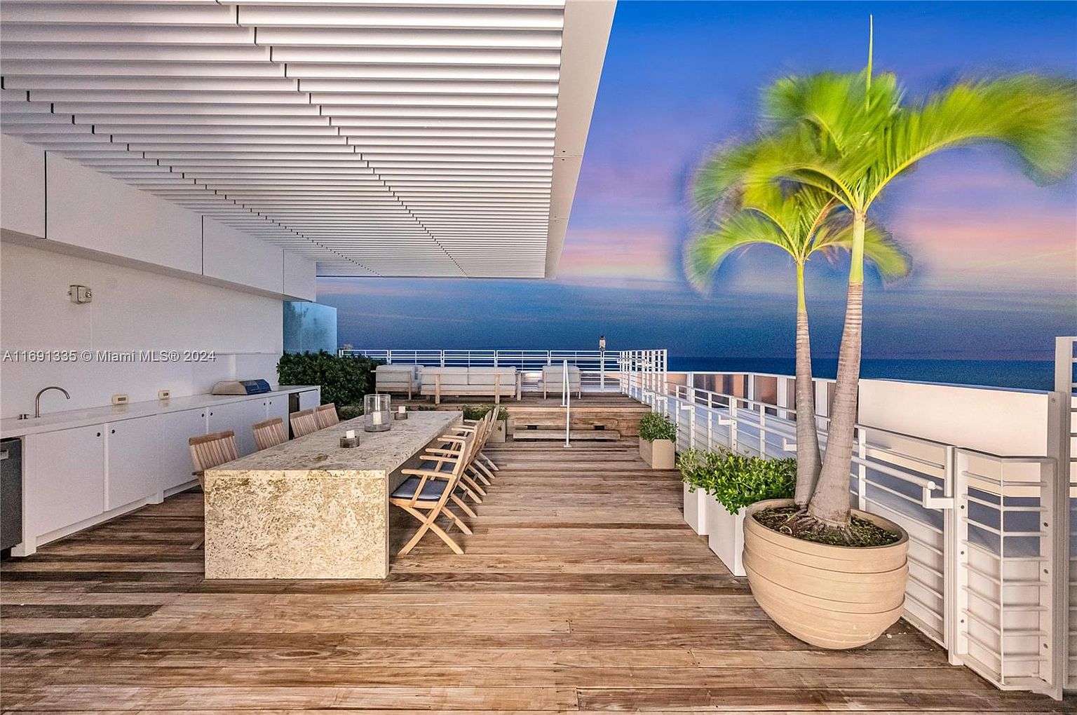 This image showcases a luxurious outdoor patio or balcony area, featuring a large dining table with seating, a built-in outdoor kitchen, and potted palm trees. The space is designed with wooden decking and white railings, offering stunning ocean views at dusk. The overall impression is one of upscale outdoor living and entertaining.