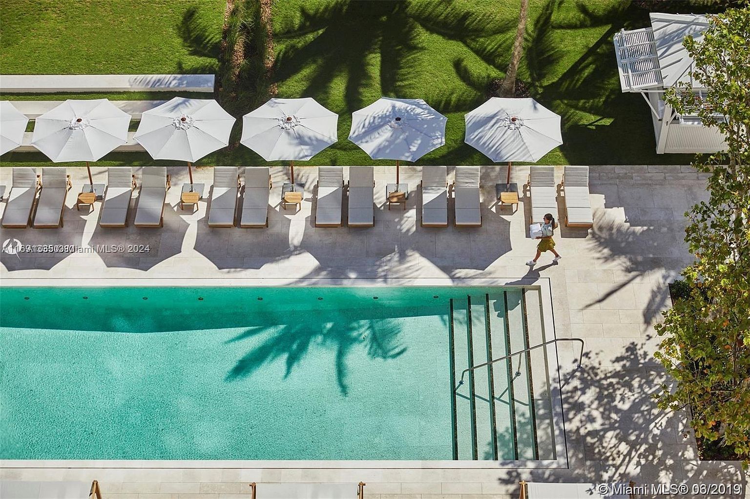 This is an aerial view of a luxurious pool area. The pool features clear turquoise water and is surrounded by a light-colored tiled deck with lounge chairs and white umbrellas. Lush green grass and palm tree shadows add to the serene and upscale ambiance, suggesting a high-end resort or residential complex.