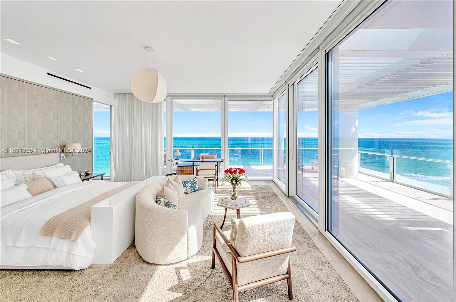 This is a bright and airy primary bedroom with stunning ocean views through large sliding glass doors that lead to a balcony. The room features a large bed, a curved sofa, and a seating area, all contributing to a luxurious and relaxing atmosphere. The neutral color palette and natural light create a serene and inviting space.