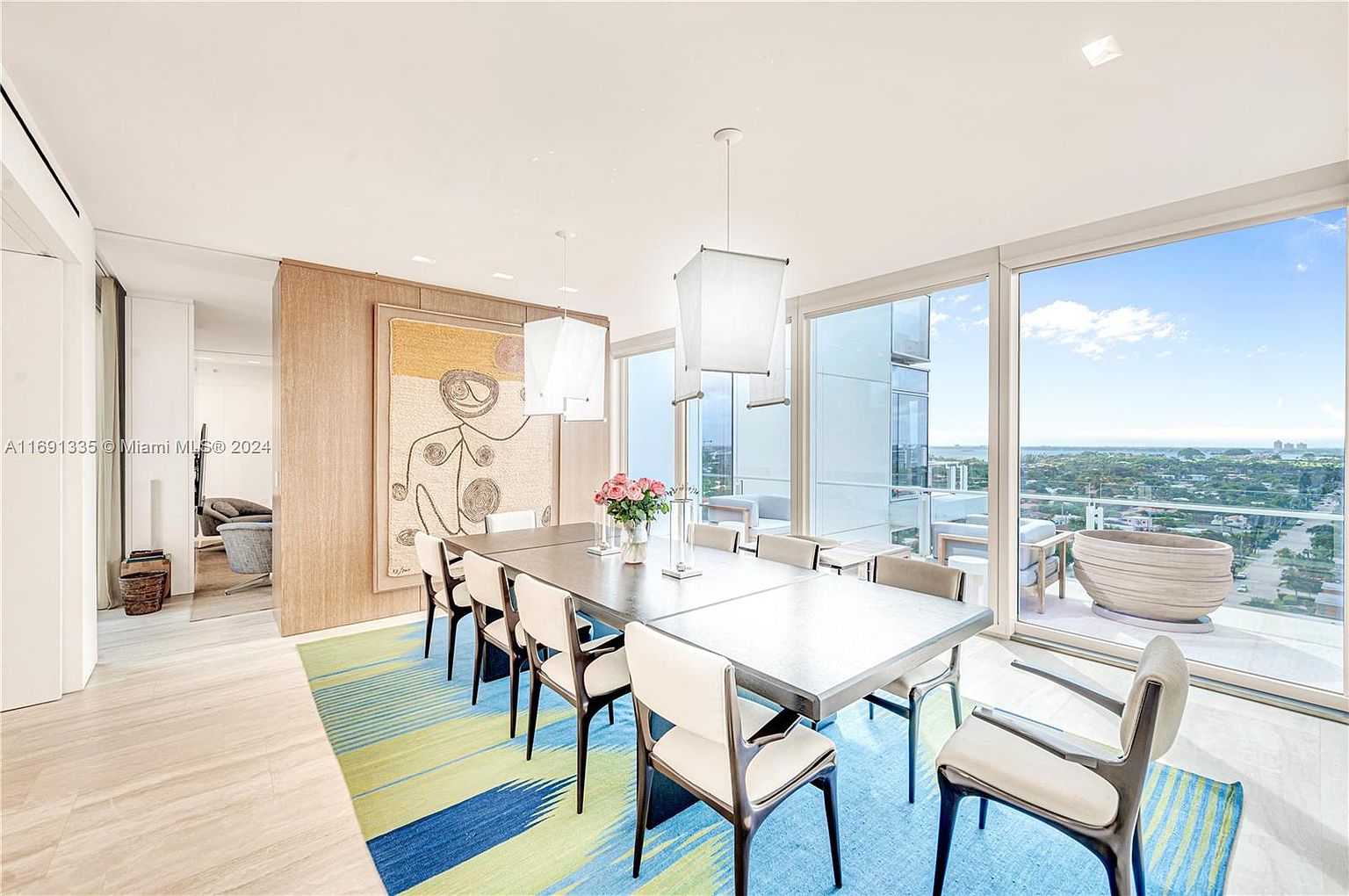 This is a bright and airy dining room featuring a large dining table with modern chairs, illuminated by contemporary pendant lighting. The room boasts floor-to-ceiling windows offering stunning views of the cityscape and ocean, leading to a balcony. An abstract art piece adds a touch of sophistication to the space, complemented by a colorful rug.