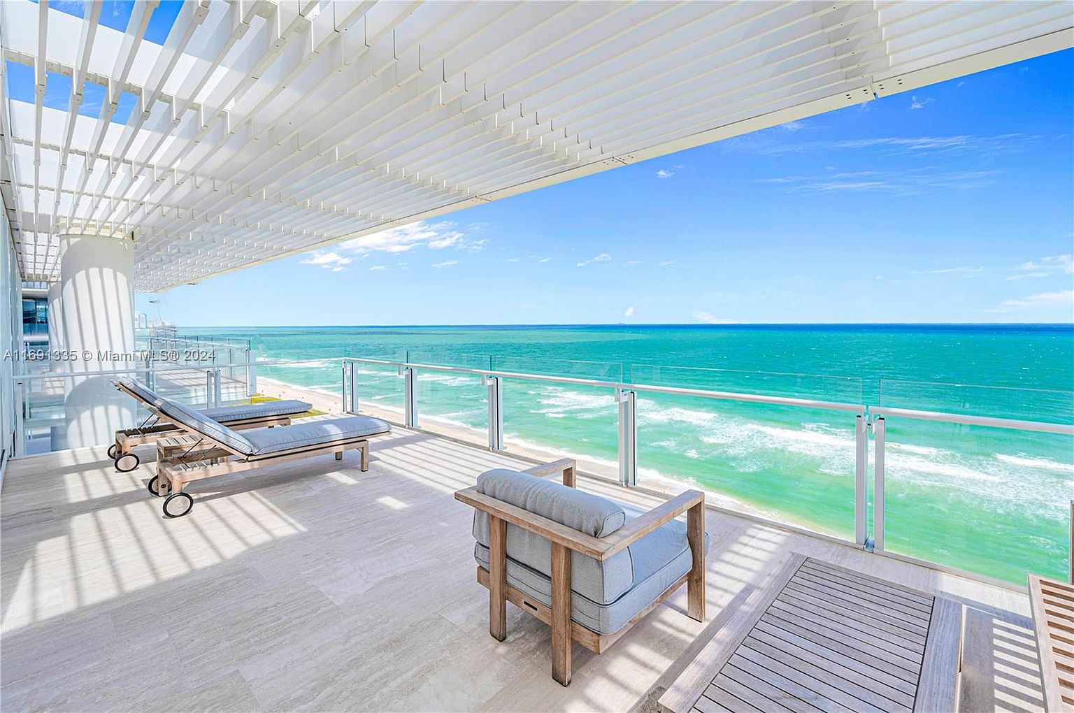 This image showcases a luxurious balcony with stunning ocean views. The space is furnished with modern outdoor furniture, including lounge chairs and a seating area, all set against a backdrop of turquoise water and a clear blue sky. A white pergola provides shade, enhancing the upscale and relaxing atmosphere of this outdoor living space.