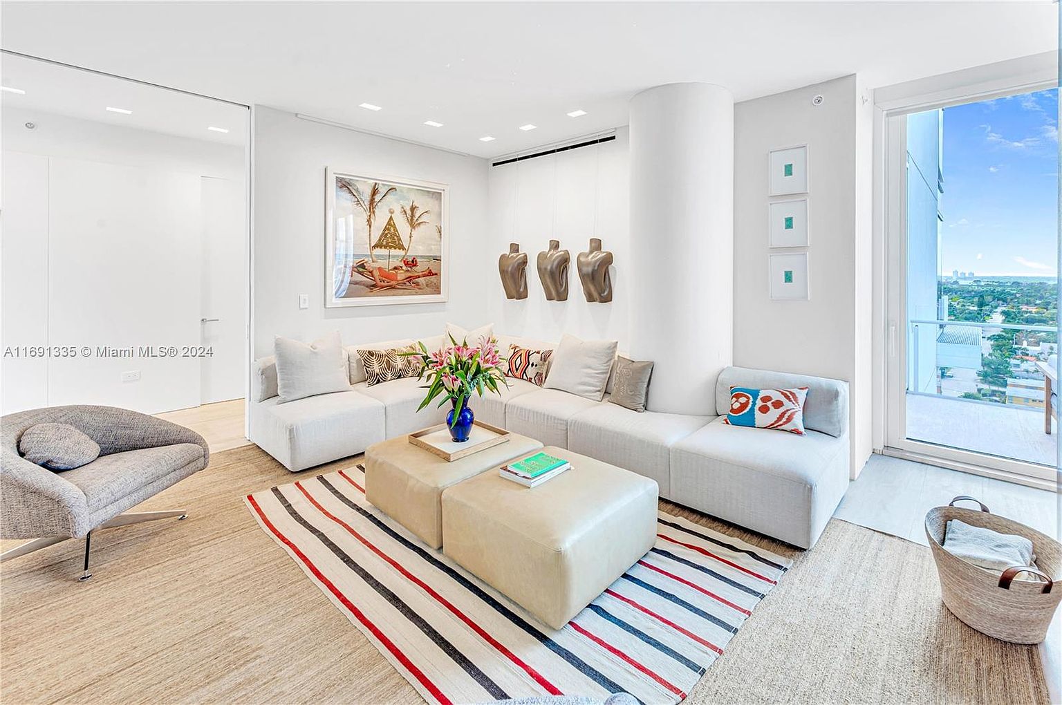 This is a bright and airy living room featuring a large sectional sofa, a pair of ottoman coffee tables, and a striped rug. The room is decorated with modern art and sculptures, and a large window offers a view of the city skyline. The overall style is contemporary and minimalist, creating a comfortable and inviting space.