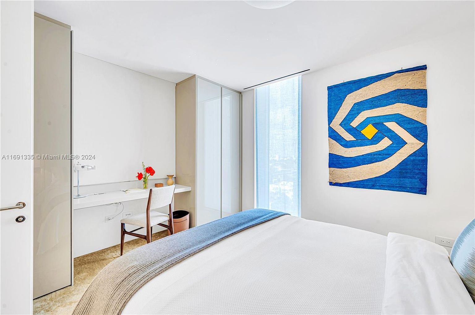 This is a bright and modern bedroom featuring a white bed with a blue and gray throw, a desk area with a white chair, and a unique blue and beige wall hanging. The room has a minimalist aesthetic with clean lines and a neutral color palette, creating a serene and inviting atmosphere. A large window with sheer curtains provides natural light.
