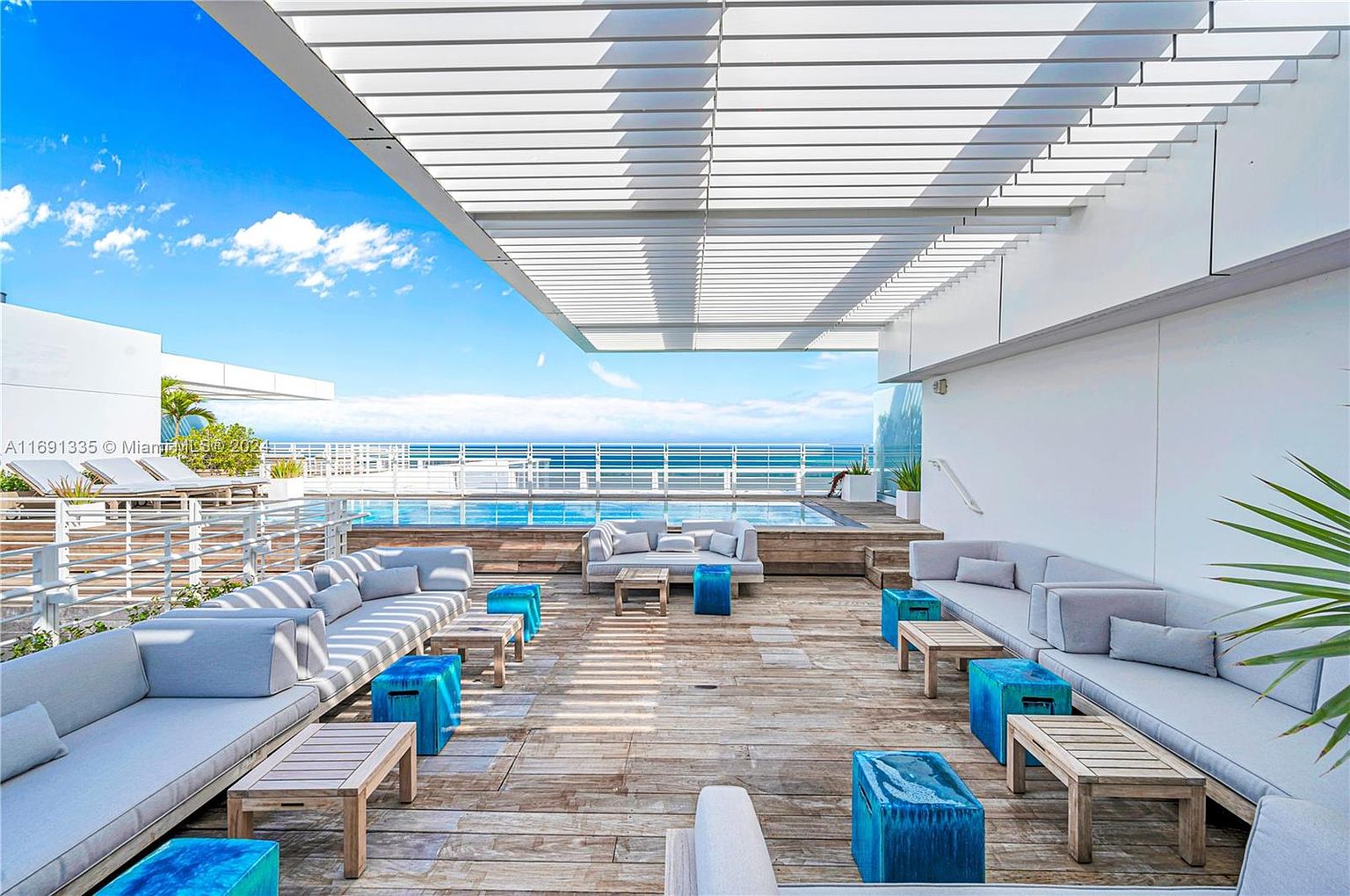 This image showcases a luxurious outdoor patio or balcony area, featuring comfortable gray lounge seating arranged around wooden coffee tables and vibrant blue accent stools. The space is partially covered by a modern pergola, providing shade while allowing glimpses of the ocean view in the background. The overall impression is one of upscale relaxation and sophisticated outdoor living.