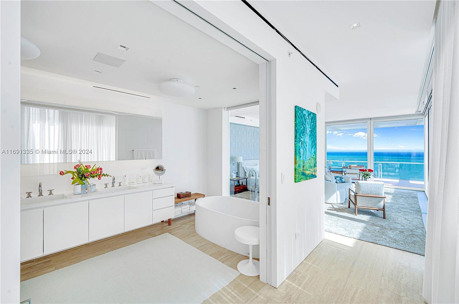 This is a bright and luxurious primary bathroom featuring a double vanity with white countertops and cabinetry, a large mirror, and a modern freestanding bathtub. The room has a clean, minimalist design with light-colored flooring and a view of the ocean through large windows, creating a spa-like atmosphere. The space flows seamlessly into an adjacent living area, enhancing the sense of openness and luxury.