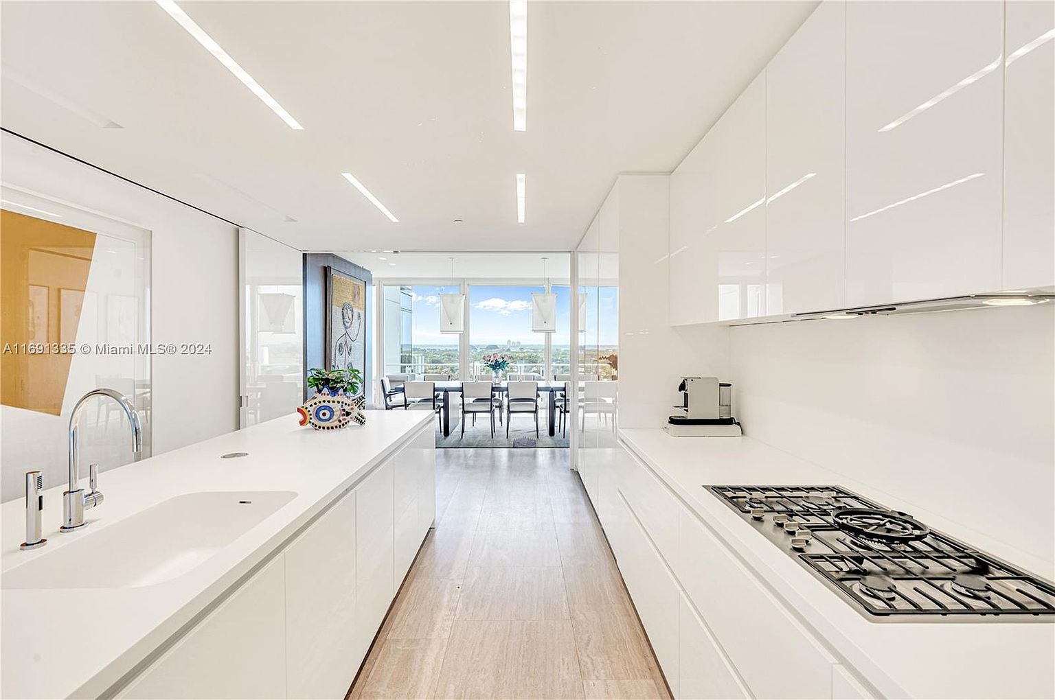 This is a bright, modern kitchen featuring sleek white cabinetry and countertops, with a gas cooktop and stainless steel appliances. The kitchen extends into a dining area with a view of the city skyline through large windows, creating an open and airy atmosphere. The perspective is a straight-on shot, emphasizing the depth and cleanliness of the space.