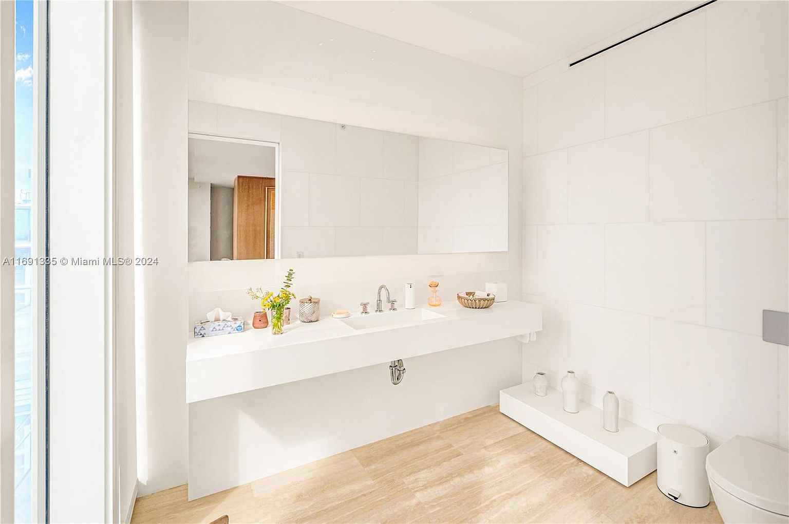 This is a bright and modern primary bathroom featuring a long, floating vanity with a white countertop and integrated sink. A large mirror spans the length of the vanity, reflecting the clean lines and minimalist design. The walls are tiled in a light color, and the flooring appears to be a light-toned stone or tile, contributing to the bathroom's airy and spacious feel.