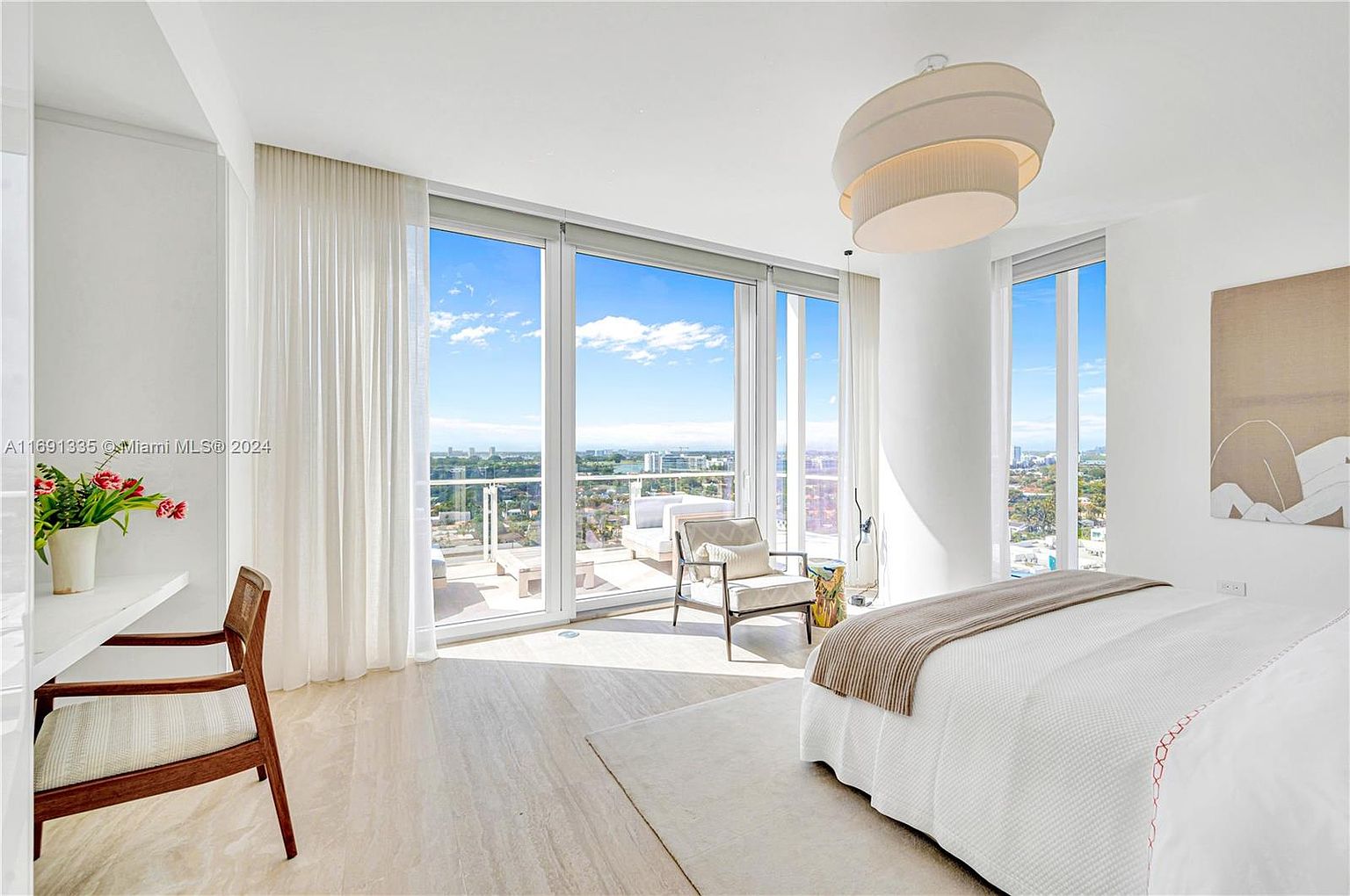 This is a bright and airy primary bedroom with floor-to-ceiling windows offering panoramic city views and access to a private balcony. The room features a neutral color palette, a large bed with a light-colored throw, and a modern drum-style light fixture. A desk and chair are positioned near the window, creating a functional workspace with a view.