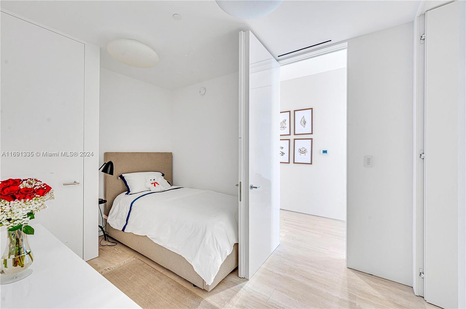This is a bright and minimalist guest bedroom featuring a twin bed with a neutral-toned headboard and white linens accented with a blue trim. The room is painted in a clean white, enhancing the sense of space and light, with a modern lamp and a small bedside table. An open doorway leads to another room, and the flooring appears to be light-colored tile or stone, contributing to the room's airy feel.