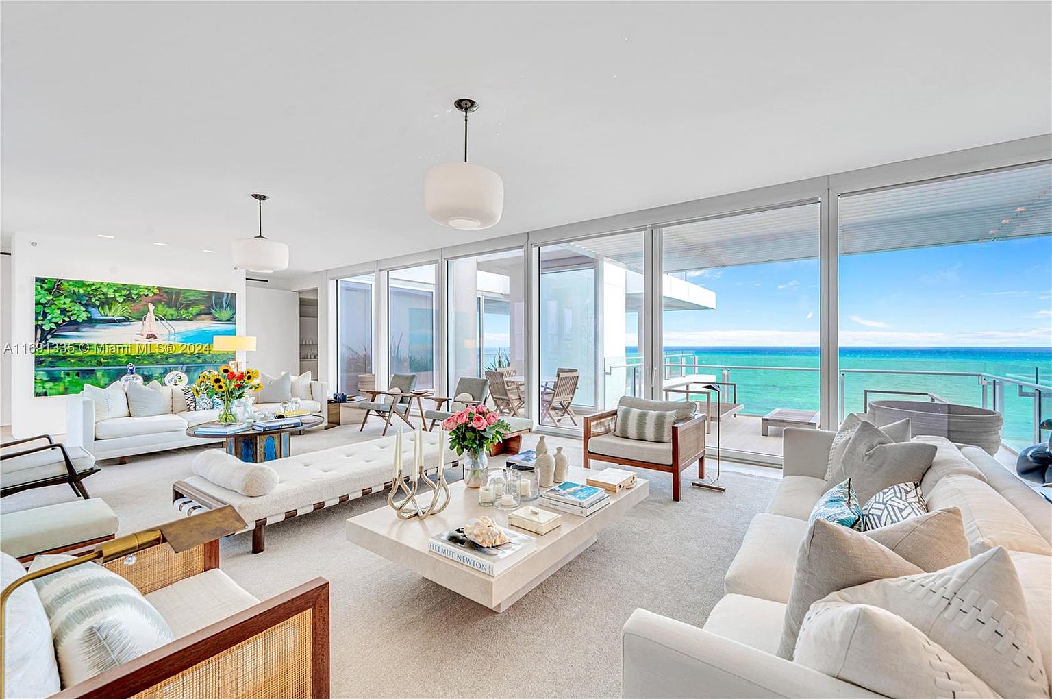 This is a bright and airy living room with floor-to-ceiling windows offering stunning ocean views. The room is furnished with white sofas, wooden-framed chairs, and a large, light-colored coffee table. The decor is modern and minimalist, creating a luxurious and inviting space.