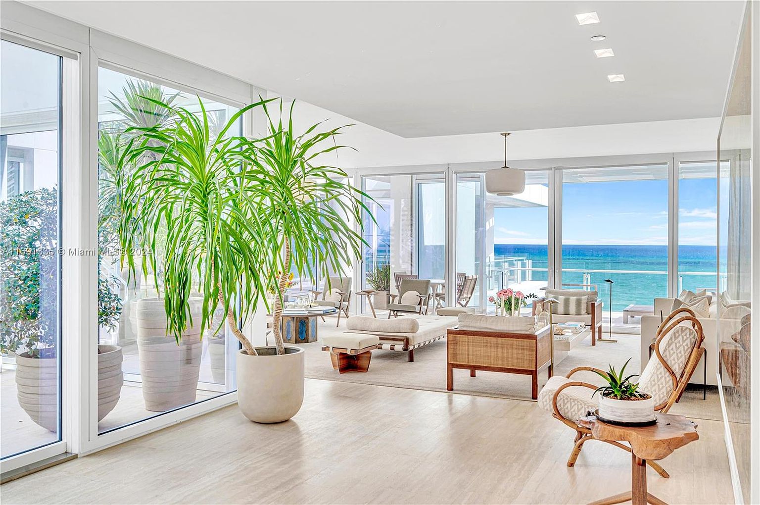 This is an interior shot of a bright and airy living room with floor-to-ceiling windows offering ocean views. The room features modern furniture, including a rattan chair and a daybed, complemented by large potted plants that bring the outdoors in. The overall aesthetic is clean, luxurious, and designed to maximize the natural light and scenic views.