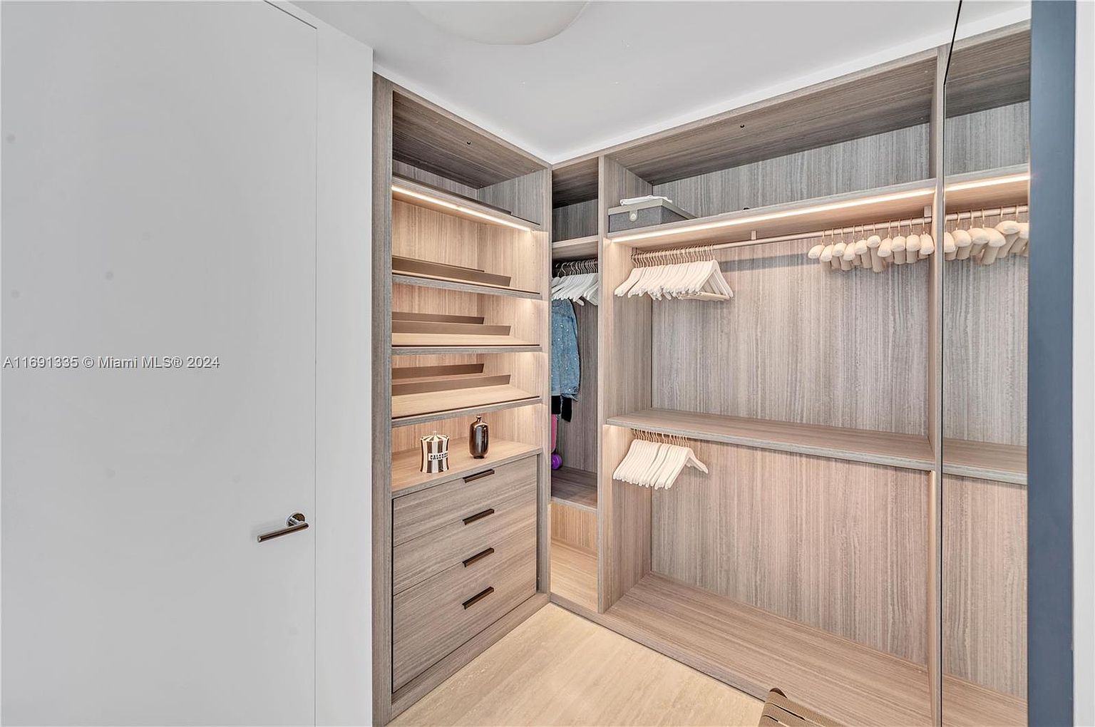 This is an interior shot of a well-organized walk-in closet. The closet features custom shelving, drawers, and hanging rods, all finished in a light wood grain. The space is illuminated by integrated lighting, creating a bright and inviting atmosphere, perfect for showcasing clothing and accessories.