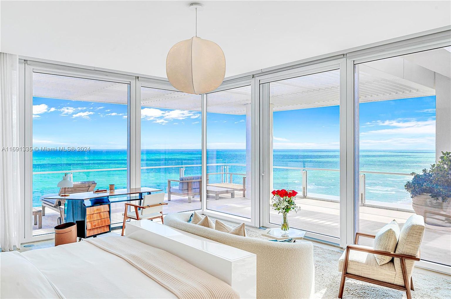 This primary bedroom boasts stunning ocean views through expansive floor-to-ceiling windows, creating a bright and airy atmosphere. The room features a neutral color palette, modern furnishings, and a minimalist design, emphasizing the serene coastal setting. A large, round pendant light adds a touch of elegance to the space.